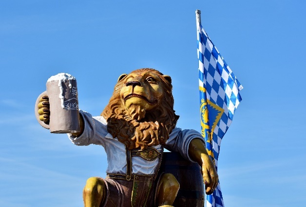 Hofrbräuhaus Löwe mit einem gefüllten Krug in der Hand, hinter ihm ein leeres Fass einer Fahne vpm Hofbräu.